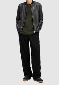 Svart läderbomberjacka över en grön texturerad stickad tröja, parat med lösa svarta byxor och svarta sneakers med bruna sulor.