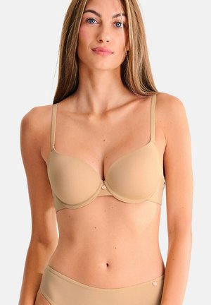 Femme aux longs cheveux portant un soutien-gorge beige rembourré et une culotte assortie, debout devant un fond clair uni.