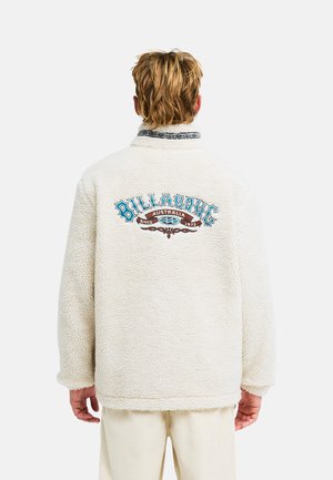 Persona che indossa una giacca in pile color crema con il logo "Billabong Australia Since 1973" in blu e marrone sulla schiena, di spalle.