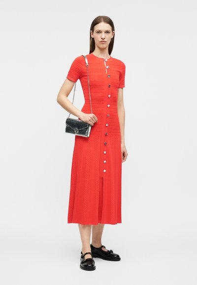 Γυναίκα που φοράει midi φόρεμα σε κόκκινο χρώμα με κουμπιά, κοντά μανίκια, μαύρα λουστρίνια παπούτσια και κρατά μια μικρή μαύρη τσάντα ώμου με αλυσίδα.