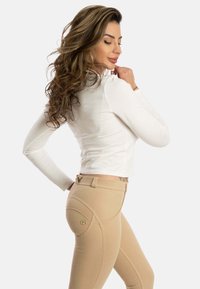 Pitkähihaisesta valkoisesta paidasta, joka on yhdistetty istuviin beigeihin ratsastushousuihin, joissa on kaarimuotoiset saumat ja pieni logo takataskussa.