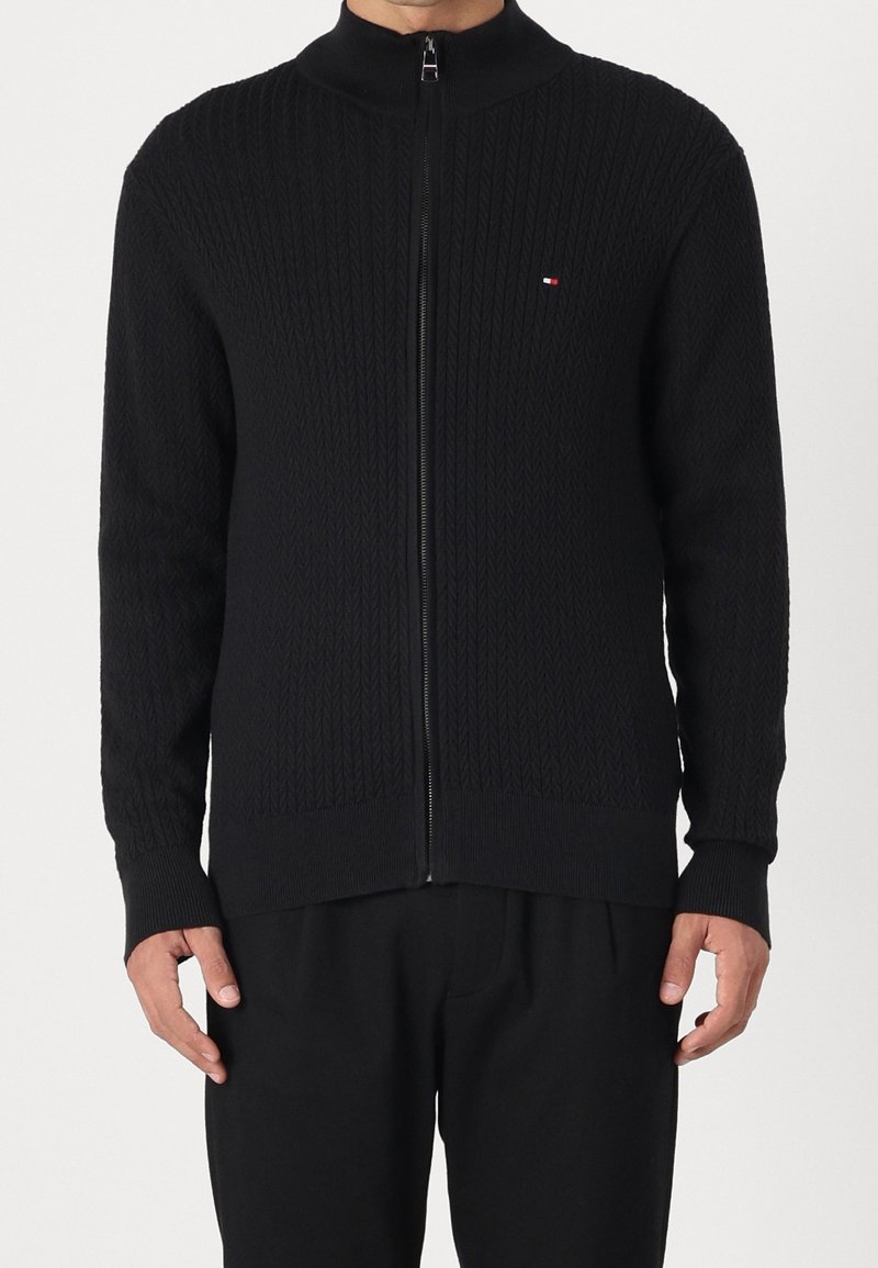 Black zip-up sweater with a textured diamond pattern, high collar, and ribbed cuffs. Features a small logo on the left chest.