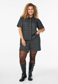 Žena s kovrdžavom kosom nosi crno-bijelu karo košulju kratkih rukava i odgovarajuće kratke hlače, crne tajice i crne čizme do koljena, s osmijehom.