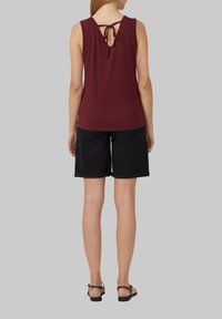 Bordeaux kleurige mouwloze top met een V-vormige rug en strikdetail, gecombineerd met zwarte shorts met opgerolde zoom, tegen een effen grijze achtergrond.