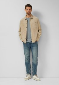 Beige Cordjacke aus Cord mit Fronttaschen, kombiniert mit einem grauen T-Shirt, hellblauen Jeans und weißen Sneakers mit beigen Akzenten.