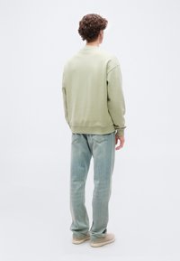 Άνδρας με σγουρά μαλλιά, φορώντας ανοιχτό πράσινο φούτερ, ανοιχτόχρωμο μπλε τζιν και μπεζ παπούτσια, στέκεται πλάτη σε απλό φόντο.