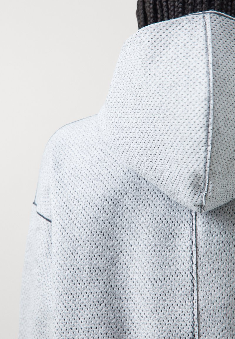 Hellblaue strukturierte Kapuzenjacke mit hohem Kragen und Kapuze. Sie verfügt über kontrastierende Nähte und ein atmungsaktives, perforiertes Stoffdesign.