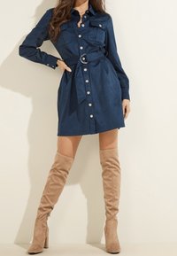 Granatowa sukienka koszulowa z guzikami, z dwoma kieszeniami na piersi, paskiem w talii i długimi rękawami, zestawiona z beżowymi zamszowymi kozakami do kolan.