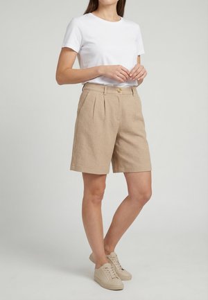 Donna che indossa una maglietta bianca a maniche corte, pantaloncini beige testurizzati fino al ginocchio con bottoni e sneakers beige con lacci, in piedi su uno sfondo semplice.