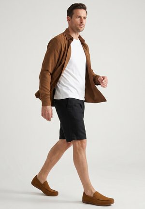Mand gående iført brun jakke, hvid t-shirt, sorte shorts og brune slip-on sko mod ensfarvet baggrund.