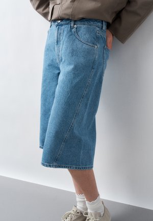 Person iført en mid-lang blå denimnederdel, hvide sokker med bølgekant, off-white sneakers og brun jakke med hænderne i lommerne.