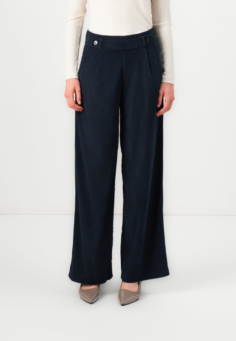Femme portant un pantalon en velours côtelé bleu foncé taille haute, à jambes larges, avec des boutons sur la ceinture, associé à des escarpins beiges à bout pointu.