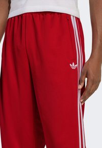 Rote Mesh-Sporthose mit weißen Seitenstreifen und einem Adidas-Logo. Verfügt über einen elastischen Bund und eine Tasche mit einem Reißverschluss.