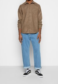 Brunukkan ruudullinen nappipaita, vaaleat siniset löysät farkut, musta-valkoiset nauhalliset tennarit, esillä neutraalilla taustalla.