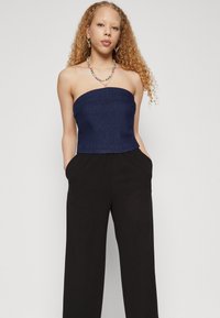 Haut en denim bleu marine sans bretelles associé à un pantalon large noir. Le mannequin porte un collier coloré en perles et des boucles d'oreilles créoles, mettant en valeur un design lisse et épuré.