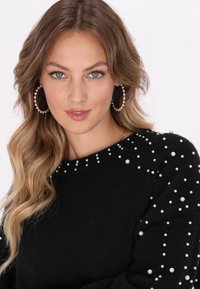 Pulover negru cu accente din perle pe umeri și guler. Modelul are părul ondulat și poartă cercei mari rotunzi. Textură netedă, design mulat.