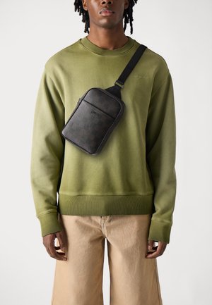 Black patterned crossbody bag made of textured material, worn over an olive green sweatshirt with ribbed cuffs, paired with beige shorts.