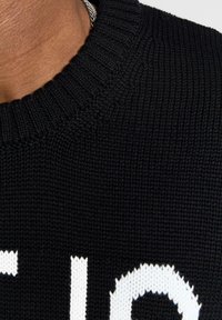 Maglione nero a maglia con scollatura a coste e un ampio motivo testuale bianco nella parte frontale. Primo piano della trama del tessuto.