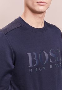 Námořnická mikina s kulatým výstřihem, s texturovaným designem na ramenou a lesklým logem v modro-černé kombinaci. Měkká textilní textura.