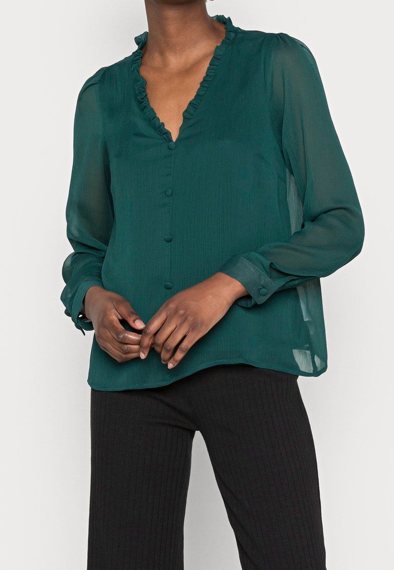 Femme portant un chemisier transparent vert foncé à manches longues avec un col en V à volants et des boutons recouverts de tissu, associé à un pantalon noir côtelé.