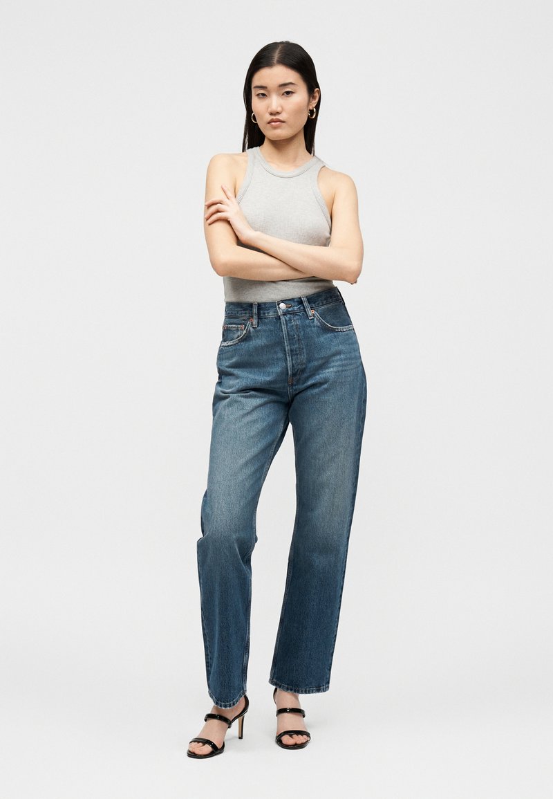 Frau mit schwarzen glatten Haaren, die ein graues ärmelloses Oberteil, blaue hochgeschnittene Jeans, schwarze Sandalen mit Absatz und goldene Creolen trägt, steht mit verschränkten Armen.