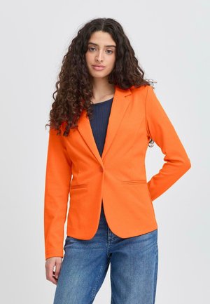 Frau mit langen, lockigen Haaren, die einen leuchtend orangefarbenen Blazer über einem dunklen Oberteil und blauen Jeans trägt und mit einer Hand in der Hüfte posiert.