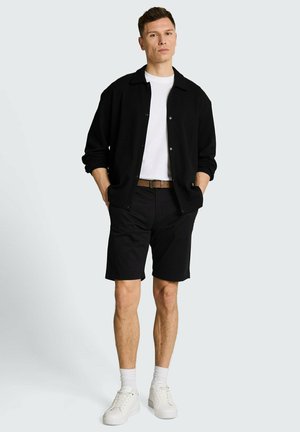 Mężczyzna stojący z rękami w kieszeniach, ubrany w czarną kurtkę, białą koszulkę, czarne krótkie spodenki, brązowy pasek, białe skarpetki i białe trampki.