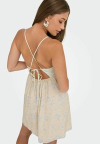 Kvinde iført en ærmeløs beige kjole med bl�å blomsterprint og krydsede bånd bagpå, set fra ryggen på en hvid baggrund.