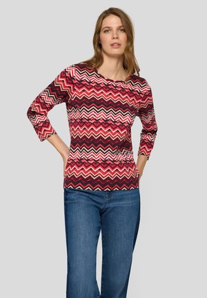 Red and pink chevron-patterned top with three-quarter sleeves, round neckline, and ribbed texture, paired with blue denim jeans.