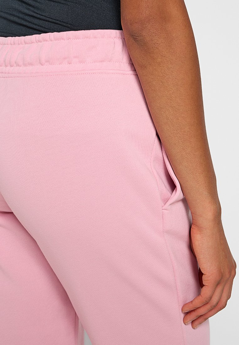 Zbliżenie na osobę w różowych dresowych spodniach z elastycznym pasem i czarnej bluzce, ręka oparta blisko bocznej kieszeni.