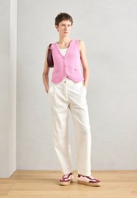Gilet rosa lavorato a maglia con scollatura a V, dotato di bottoni e tasche, abbinato a pantaloni larghi color crema e sneakers rosse e rosa.