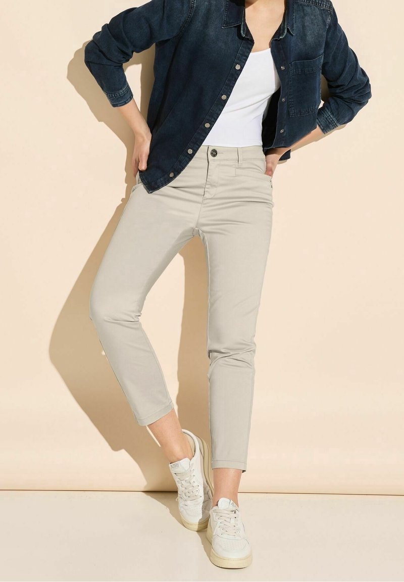 Beige ankelkorta byxor med slim fit, kombinerat med en mörk denimjacka och vita sneakers. Outfiten har en enkel vit topp.