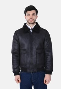 Giovane uomo che indossa una giacca di pelle nera con collo in shearling, jeans blu e una camicia bianca, in piedi davanti a uno sfondo neutro.