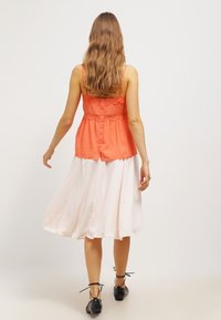 Orange ärmlös topp med spetsdetaljer och knappstängning bak, ihop med en flödande, knälång vit kjol. Svarta platta skor.