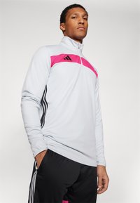 Haut de sport à manches longues gris clair avec une bande rose et des accents noirs ; il présente une fermeture éclair quart et trois bandes verticales noires sur les côtés.