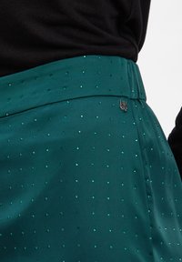 Turkusowa spódnica z małymi zielonymi cyrkoniami i metką z logo, noszona z czarną prążkowaną bluzką, podkreślającą talię i biodra.