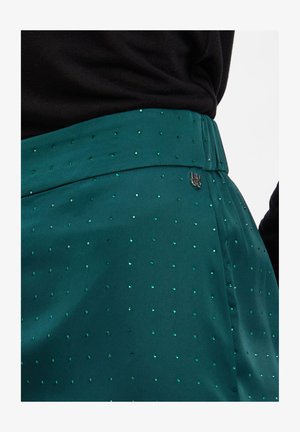 Türkise Rock mit kleinen grünen Strasssteinen und einem Logo-Tag, getragen mit einem schwarzen Rippoberteil, das die Taille und Hüften betont.