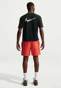 Άνδρας που στέκεται με την πλάτη, φοράει μαύρο t-shirt Nike με μεγάλο λευκό σύμβολο, κόκκινα σορτς, λευκές κάλτσες και μαύρα αθλητικά παπούτσια με πορτοκαλί λεπτομέρειες.