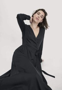 Schwarzes Wickelkleid mit langen Ärmeln aus glattem Stoff, mit einem V-Ausschnitt und einem gebundenen Taillenbereich, dazu ein fließender Rock, der nach unten ausläuft.