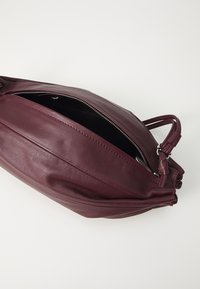 Borsa in pelle bordeaux con design angolato, chiusura a zip e due manici. L'interno è scuro con cuciture visibili all'esterno.