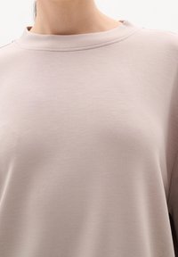 Gros plan d'une personne portant une chemise à manches longues beige clair, avec un col rond et une texture de tissu lisse.