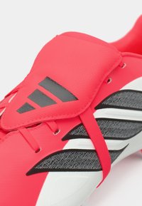 Κόκκινο ποδοσφαιρικό παπούτσι με υφή συνθετικού υλικού, μαύρες λεπτομέρειες, κομψό σχέδιο με ξεχωριστές ρίγες και ασφαλές κάλυμμα κορδονιών.