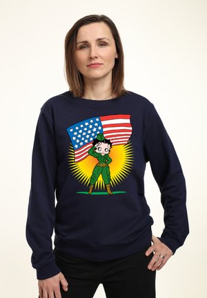 Námořnická mikina s kreslenou postavičkou v zeleném oblečení, která salutuje, na pozadí s vlajkou USA a žlutými paprsky, textura látky.