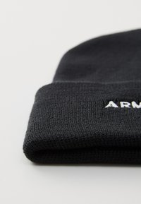 Berretto in maglia nera con risvolto, caratterizzato da testo ricamato bianco che recita "ARM" sul davanti. Tessuto testurizzato con un pattern a maglia fitta.