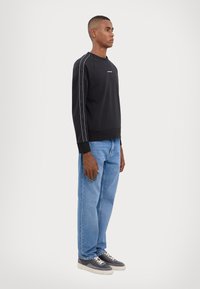 Svart bomulls hettegenser med hvite sidestiper, brukt med lyseblå jeans og grå joggesko. Enkelt design og avslappet passform.