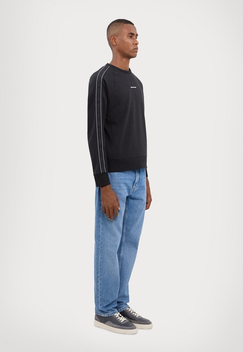 Svart bomulls hettegenser med hvite sidestiper, brukt med lyseblå jeans og grå joggesko. Enkelt design og avslappet passform.