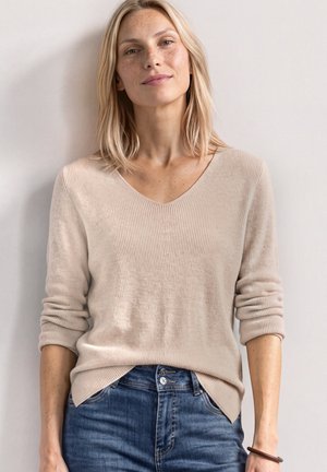 Femme blonde avec des taches de rousseur portant un pull beige à col en V et un jean bleu, debout contre un mur clair les bras détendus.