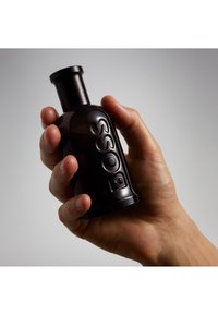 Hand hält eine kleine matte schwarze Flasche, auf deren Seite vertikal das Wort "BOSS" geprägt ist, vor einem neutralen Hintergrund.