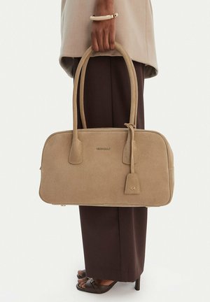Persona sosteniendo un bolso beige con doble asa, vistiendo pantalones marrones, chaqueta beige y una pulsera blanca, de pie contra un fondo liso.