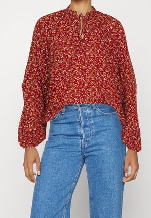 Woman wearing a red floral long-sleeve blouse with a ruffled neckline and blue high-waisted jeans, standing against a plain background.